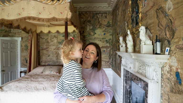 A mum holds her young daughter in her arms as they both smile looking at historic Chinese wallpaper